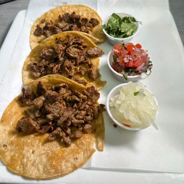 Three carne asada tacos with sides of cilantro, pico de gallo, and diced onions.