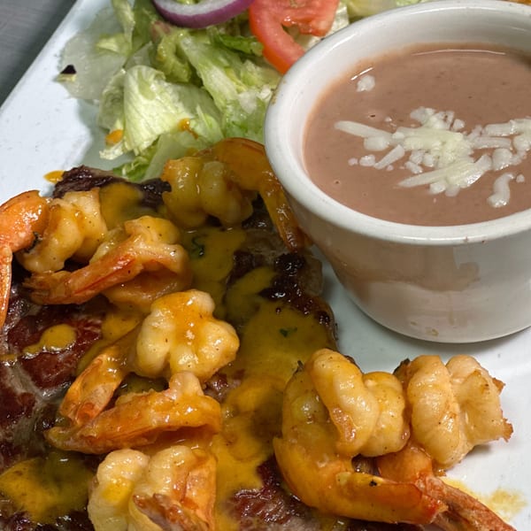 Mexican dish with shrimp and steak covered in yellow sauce, a side of refried beans, and salad.